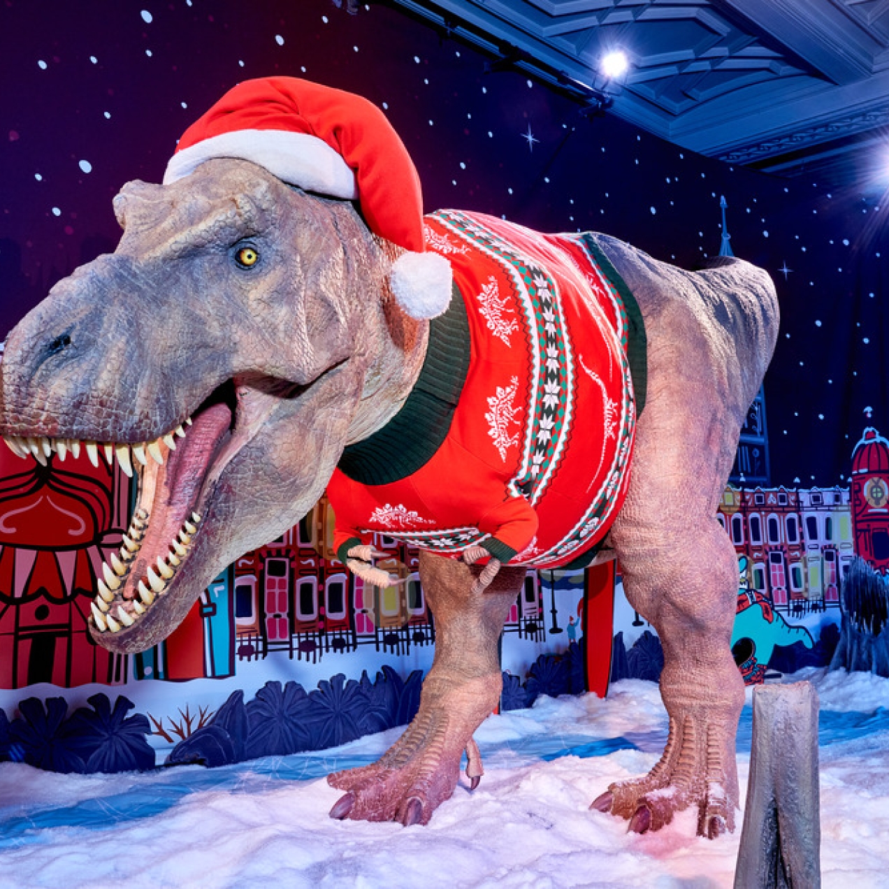 A large animatronic T-rex wearing a festive jumper and Santa hat. The background is an illustration of snow covered buildings